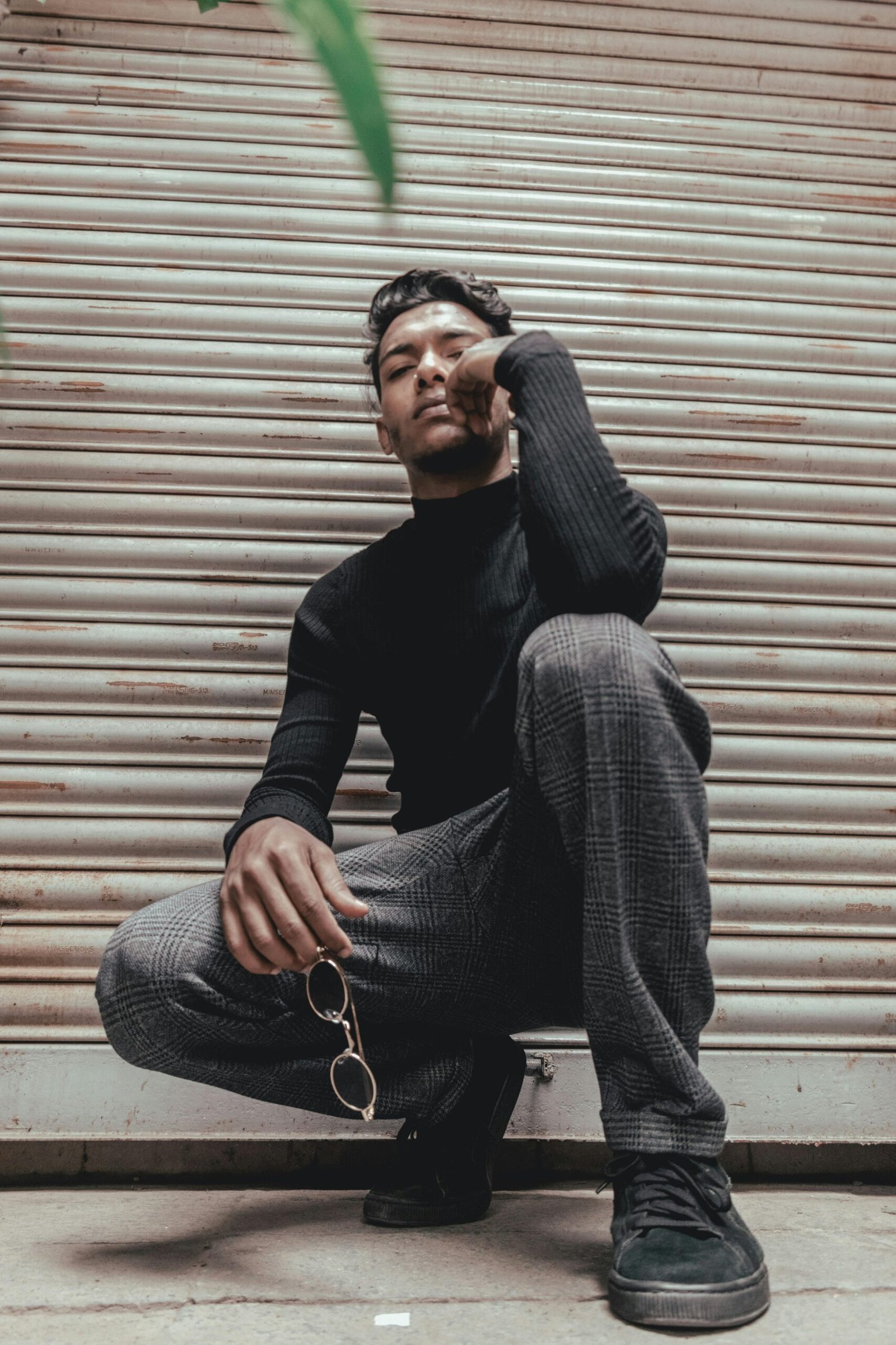 Male model posing stylishly outdoors holding sunglasses against a shutter backdrop.